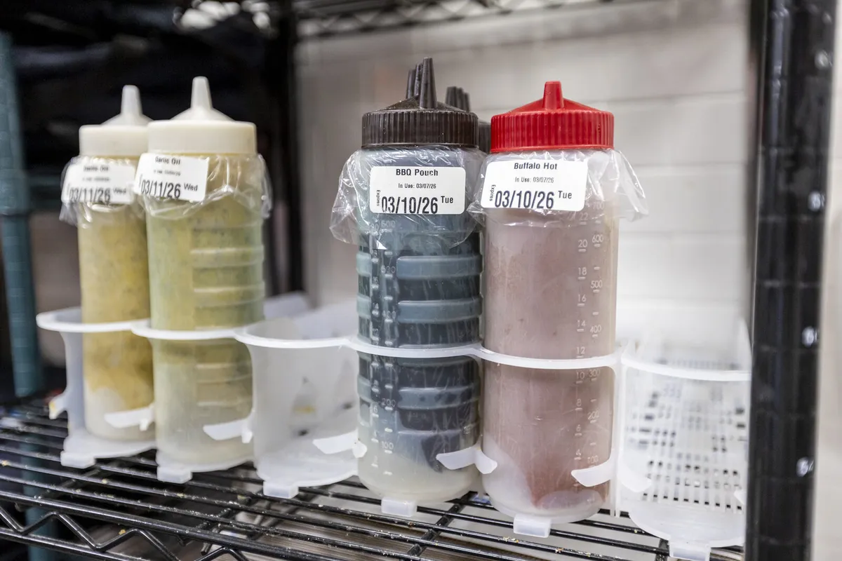 Squeeze bottles of sauce on a cooler shelf, each marked with a dated dissolvable label